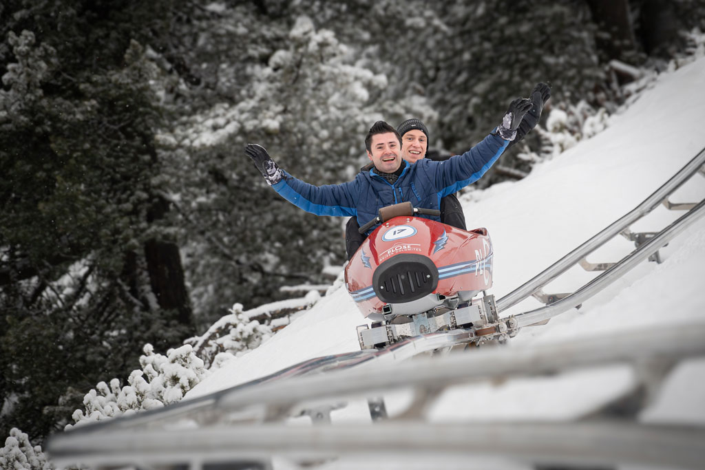 Two people riding the Plosebob on the snowy rail track of the Plose, surrounded by a winter forest.