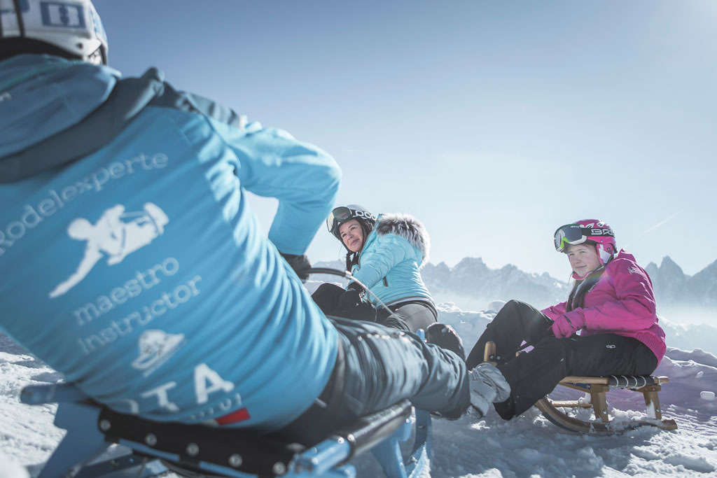 Group sledding with an instructor on the Plose, children and adults enjoying winter tobogganing with Dolomite views.